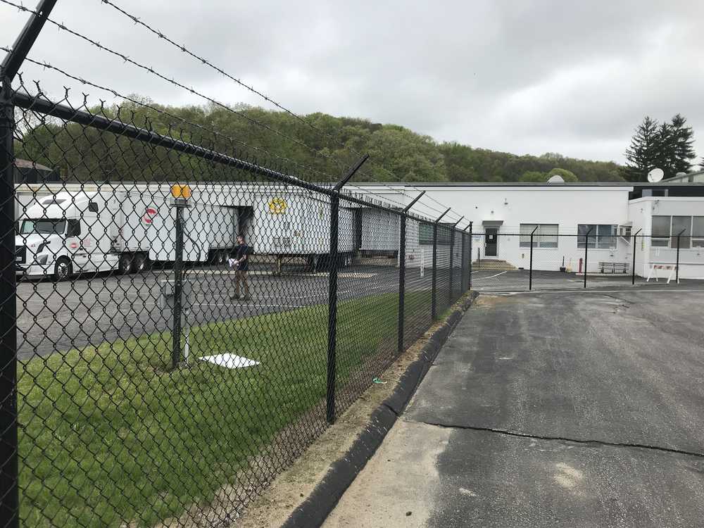 Chain-link fence surrounds a large industrial building with multiple loading docks. A truck is parked near a dock. Overcast sky and tree-covered hills in the background. Paved area and grassy strip visible.