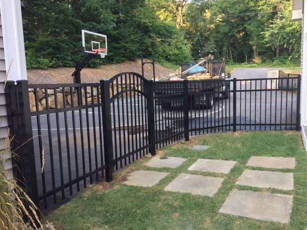A black metal fence with an arched gate encloses an outdoor area. Beyond the fence, there's a basketball hoop above a driveway, and a truck loaded with items. The area inside the fence has grass and stone tiles. Trees are in the background.