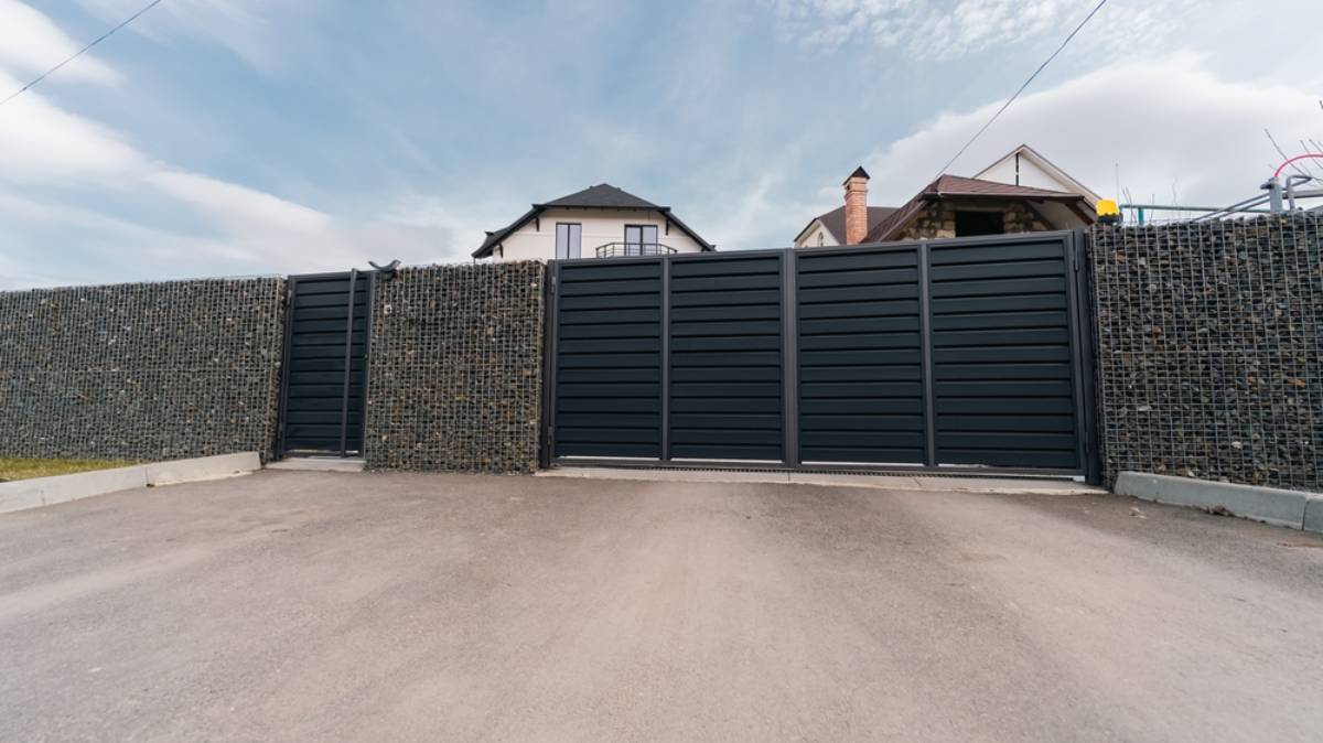 A modern black metal gate set in a stone-filled wire fence, with houses and a blue sky visible in the background. The gate is closed and the area in front is paved.