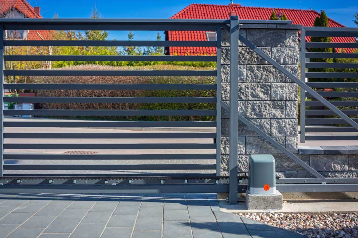 Modern gray metal sliding gate with horizontal bars and a stone pillar, partially open over a paved driveway, with houses and greenery visible in the background under a blue sky.