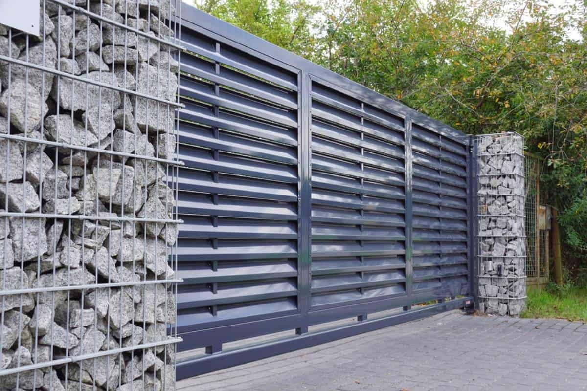 Modern metal sliding gate with horizontal slats, flanked by stone-filled wire mesh pillars. Trees and greenery are visible in the background, and the ground is paved with gray bricks.